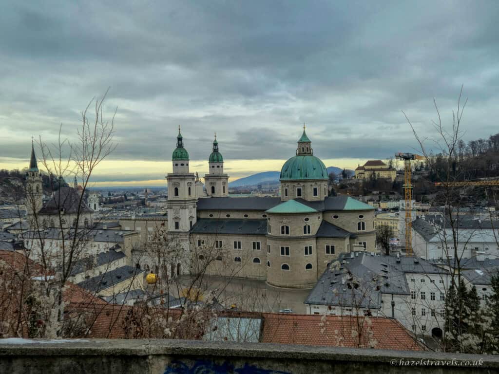 View over Salzburg with the green domes and twin towers of Salzburg Cathedral rising above historic rooftops under a cloudy sky