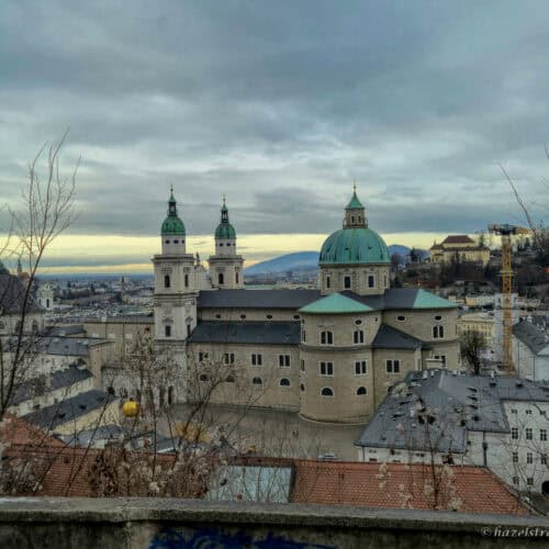 View over Salzburg with the green domes and twin towers of Salzburg Cathedral rising above historic rooftops under a cloudy sky