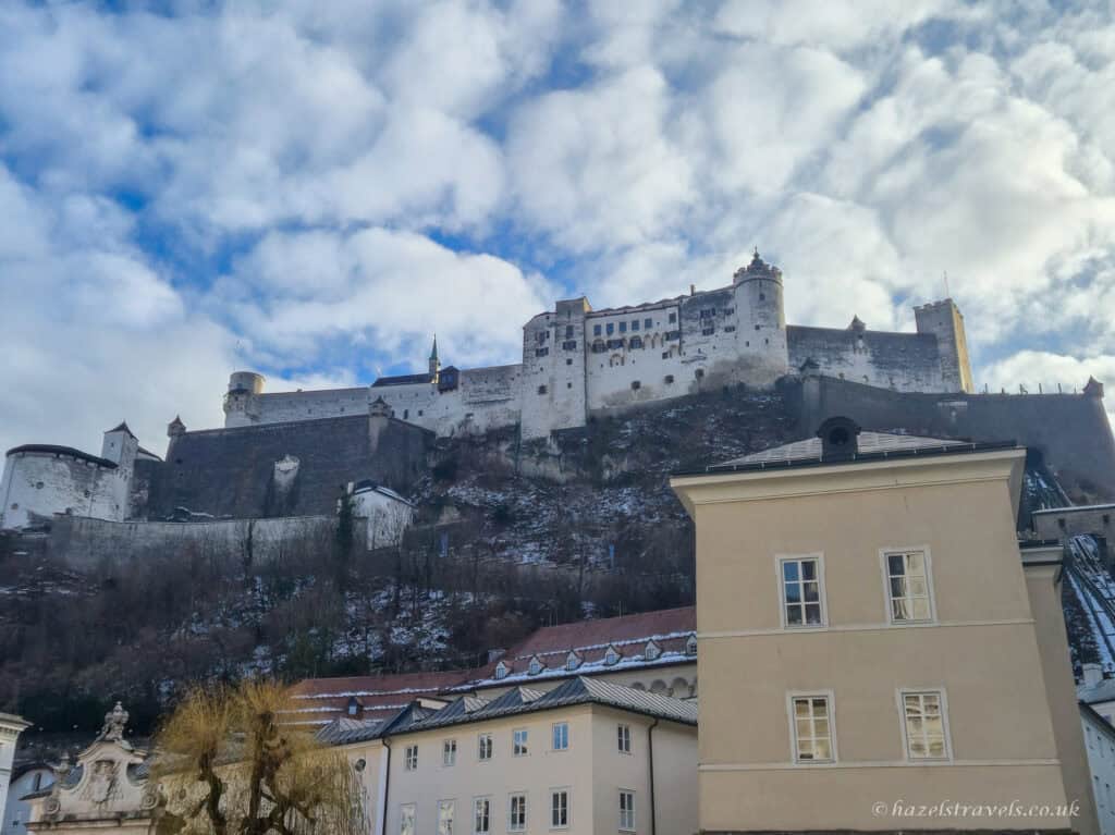 Hohensalzburg Fortress on the hill above Salzburg with white stone walls, snowy hillside, and pale historic buildings below under a cloudy blue sky