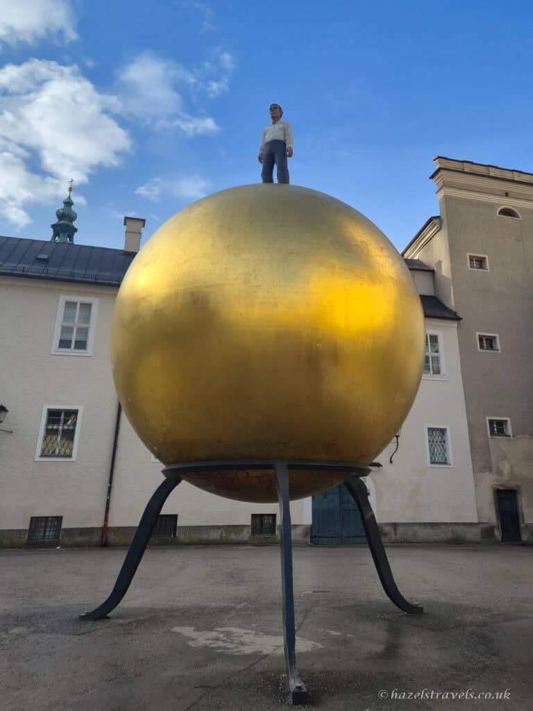 Golden Sphaera sculpture in Kapitelplatz Salzburg with a large gold sphere and a man standing on top, set against pale historic buildings and blue sky