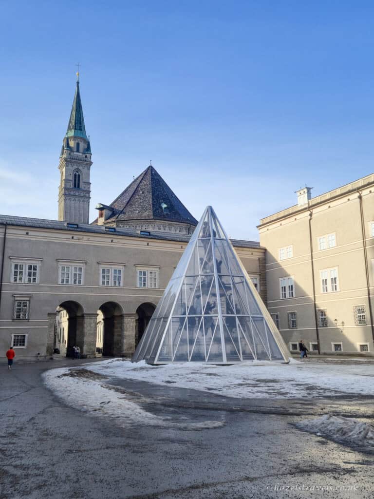 Modern glass pyramid sculpture in Kapitelplatz Salzburg with pale historic buildings, archways, and a tall church tower with green spire under a clear blue sky