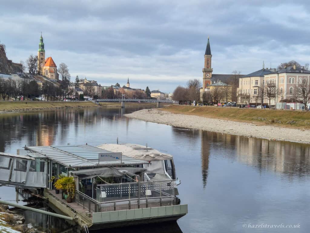 View along the Salzach River in Salzburg with calm water reflections, church towers, pastel historic buildings, and a riverside boat café under a cloudy sky