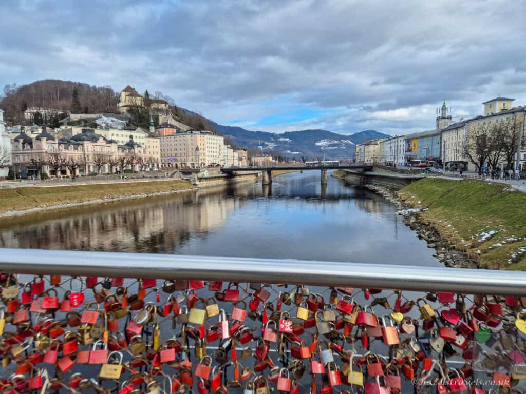 Colourful love locks attached to Makartsteg bridge railing in Salzburg overlooking the Salzach River, with historic pastel buildings, bridge, and mountains in the background