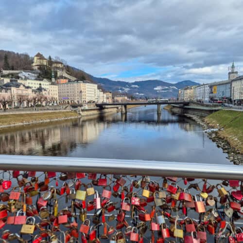 Colourful love locks attached to Makartsteg bridge railing in Salzburg overlooking the Salzach River, with historic pastel buildings, bridge, and mountains in the background
