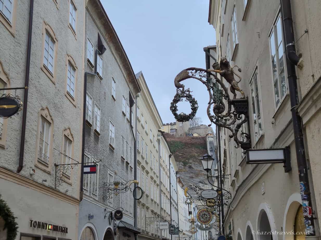 Narrow street in Salzburg Old Town with ornate wrought-iron shop signs and pastel historic buildings, with fortress walls visible on the cliff above
