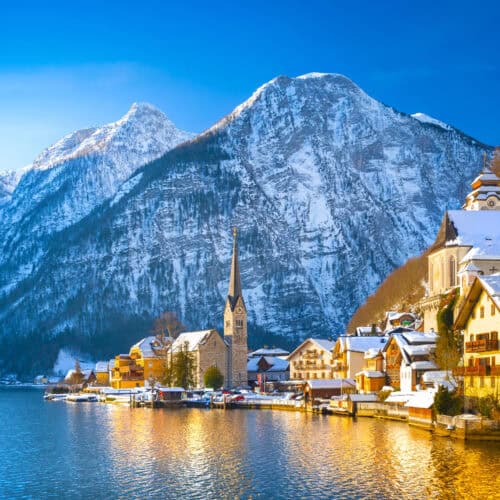 Snow-covered village of Hallstatt reflected in Lake Hallstatt with the Evangelical Church spire and dramatic Austrian Alps in the background.