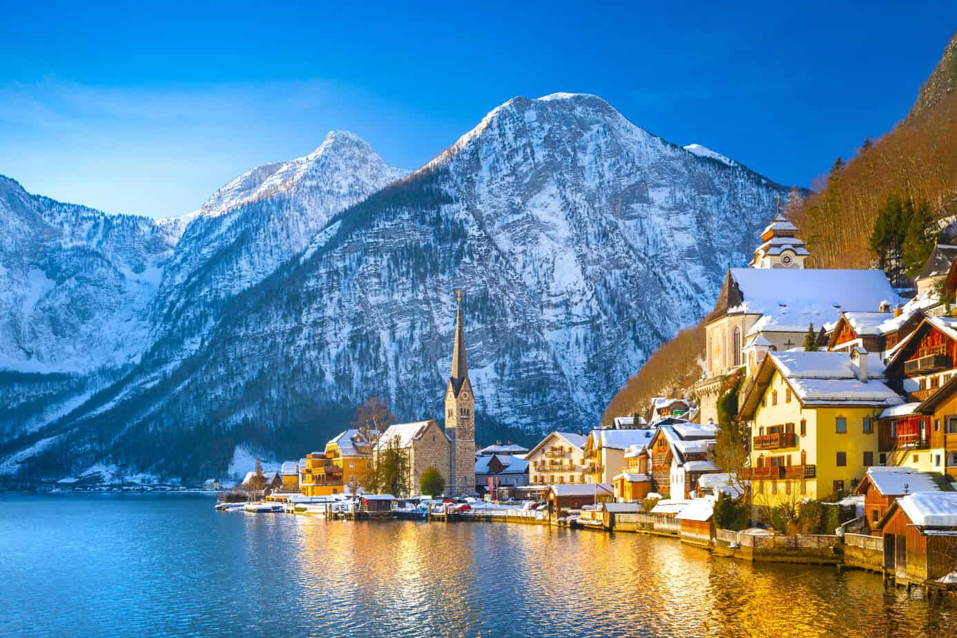 Snow-covered village of Hallstatt reflected in Lake Hallstatt with the Evangelical Church spire and dramatic Austrian Alps in the background.