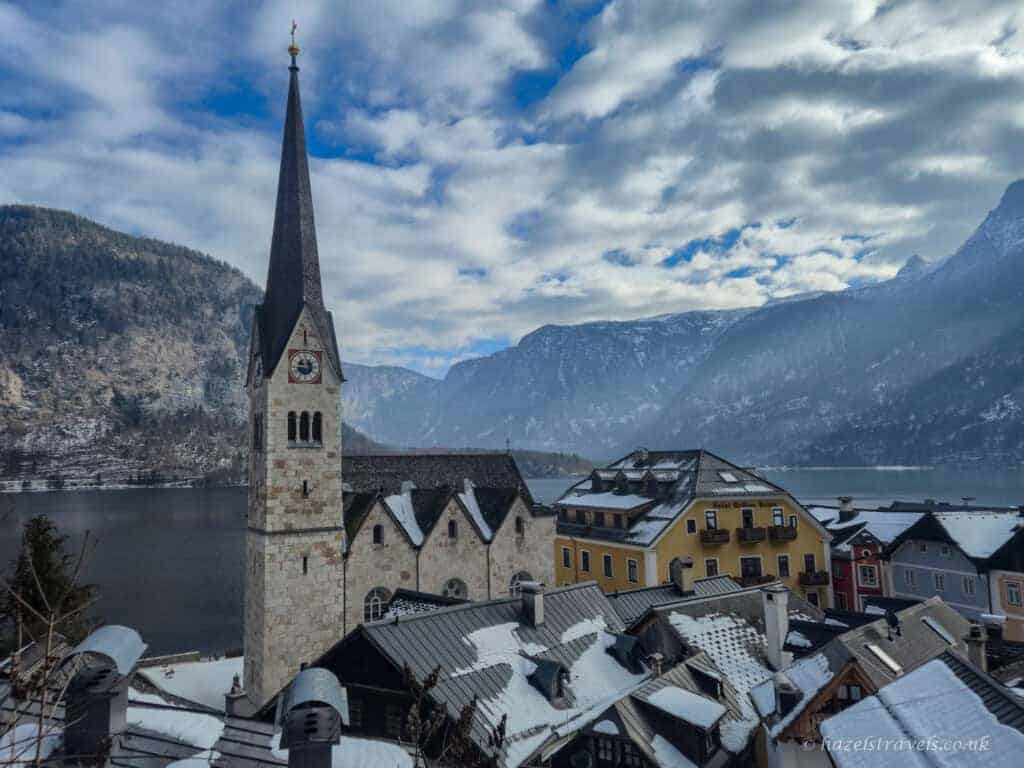 Tall spire of Hallstatt’s Evangelical Church beside Lake Hallstatt, with snow-covered rooftops and dramatic mountains under a cloudy winter sky.