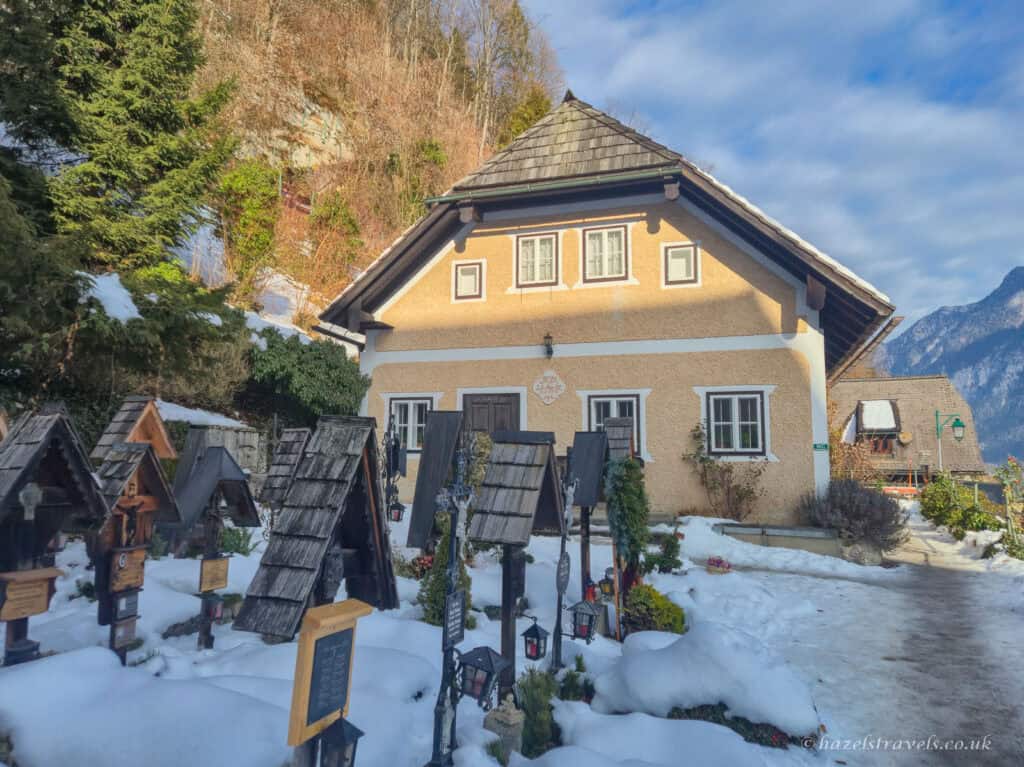 Snow-covered cemetery in Hallstatt with small wooden grave shrines in front of a pale yellow alpine house and a wooded hillside behind.