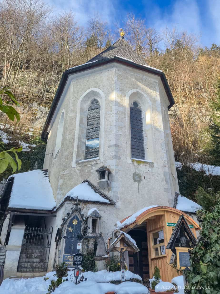 A pale stone church tower in Hallstatt rising above a snowy cemetery, with a small wooden entrance to the Bone House and snow-covered shrines at the base.