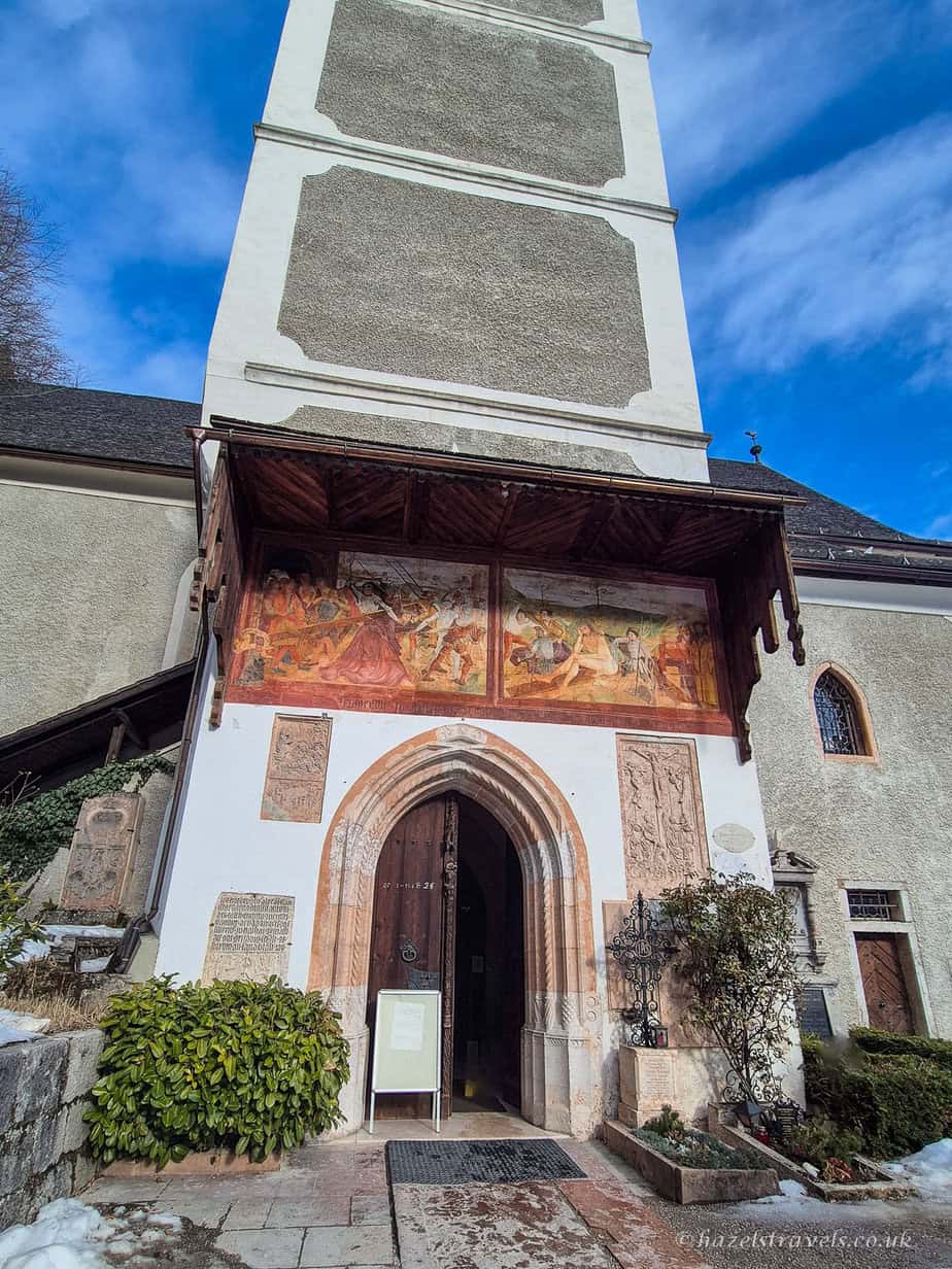 Entrance to Hallstatt’s Catholic Parish Church with an arched wooden doorway and colourful religious fresco above, beneath a tall pale church tower against a blue sky.