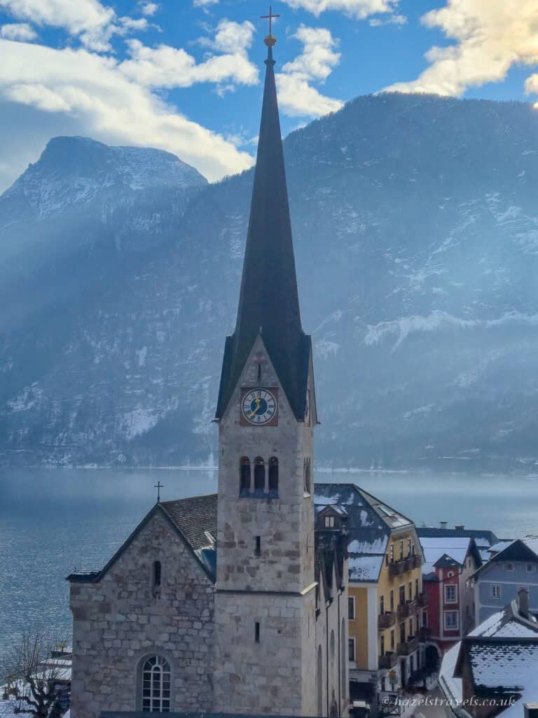 Tall dark church spire of Hallstatt’s Evangelical Church rising above stone buildings beside Lake Hallstatt, with snowy mountains and blue sky behind.