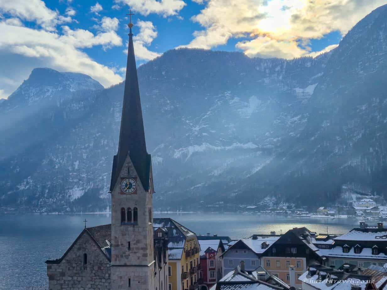 Tall stone church with a dark spire and clock tower in Hallstatt, with snow-covered village rooftops, Lake Hallstatt, and towering mountains illuminated by winter sunlight.