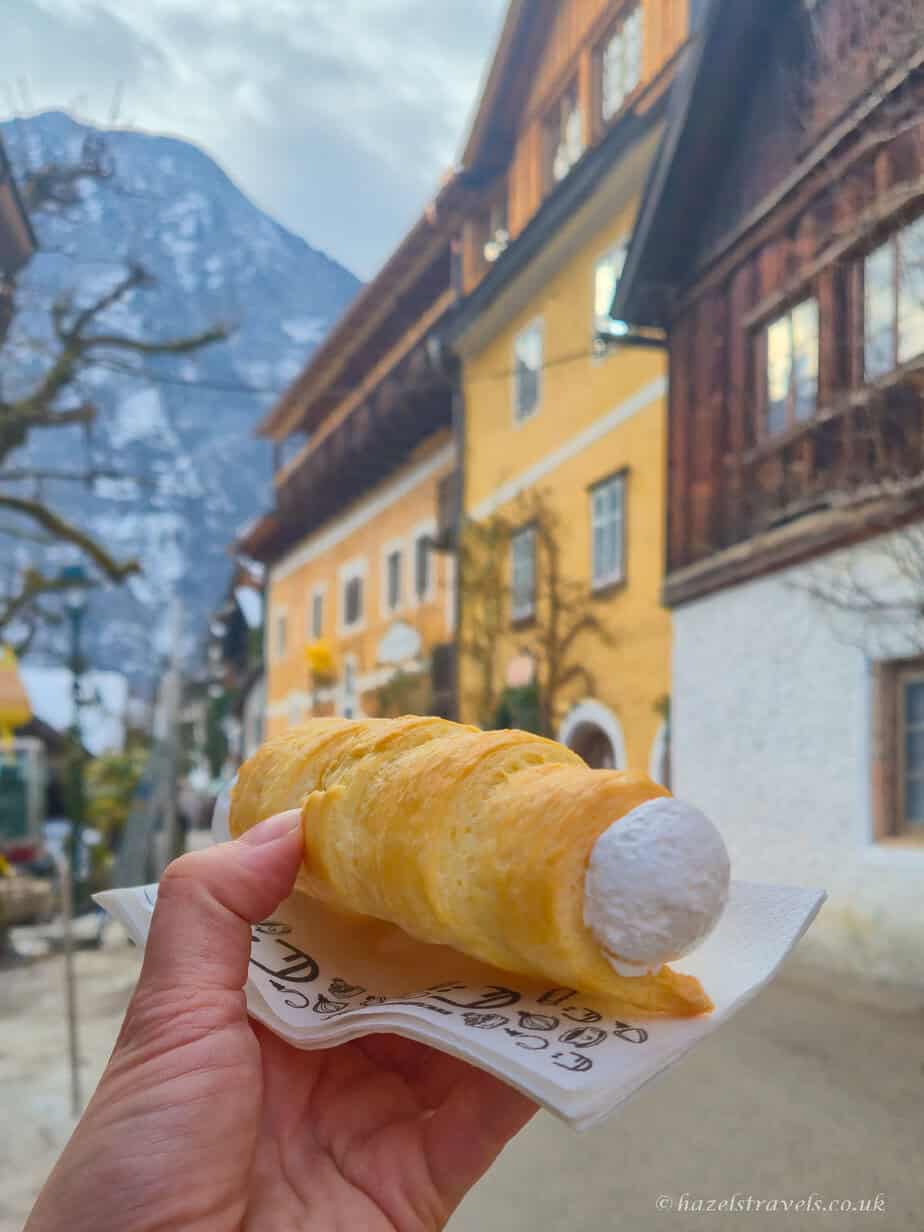 Hand holding a cream-filled pastry in Hallstatt with colourful alpine houses and snowy mountains blurred in the background.