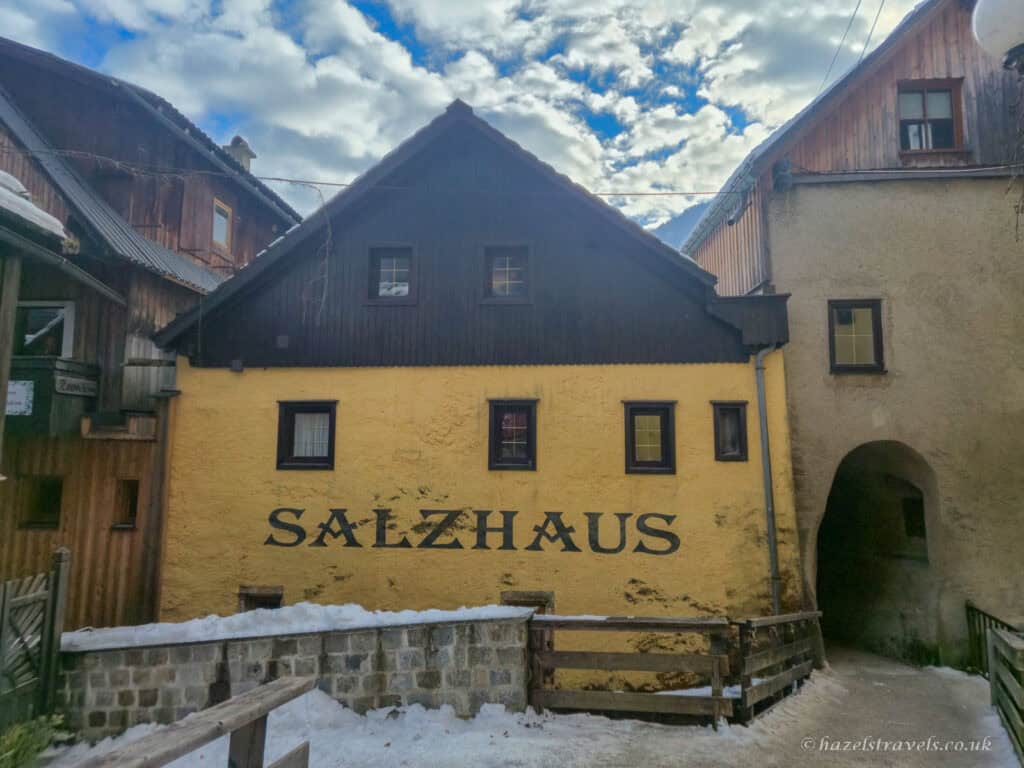 Yellow Salzhaus building in Hallstatt with the word “Salzhaus” painted across the wall, beside wooden alpine houses and a narrow archway passage, with snow on the ground.