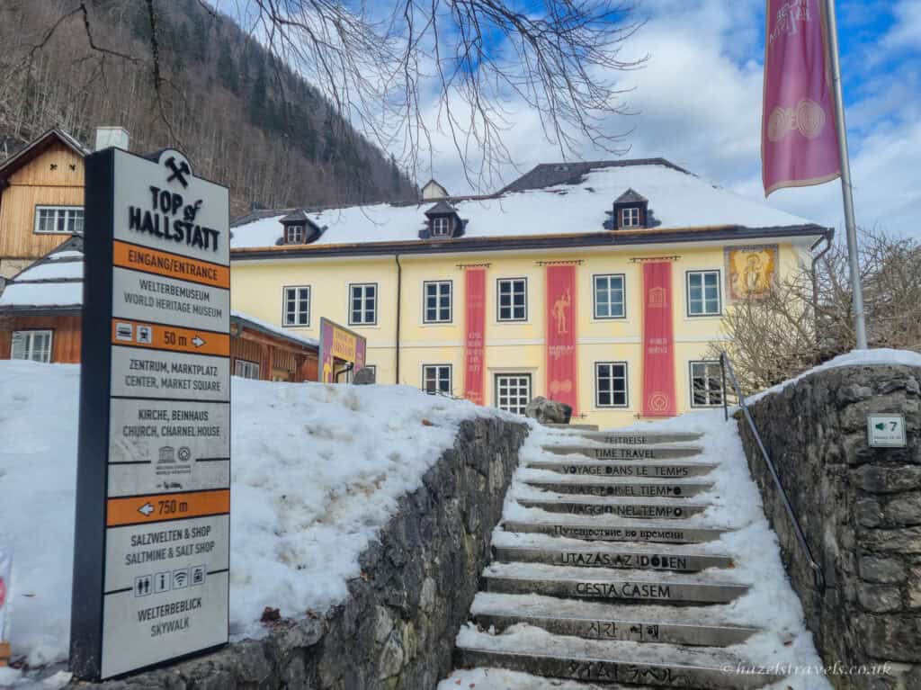 Snow-covered steps leading up to the Hallstatt World Heritage Museum building, with red banners on a yellow facade and a sign reading “Top of Hallstatt” beside the path.