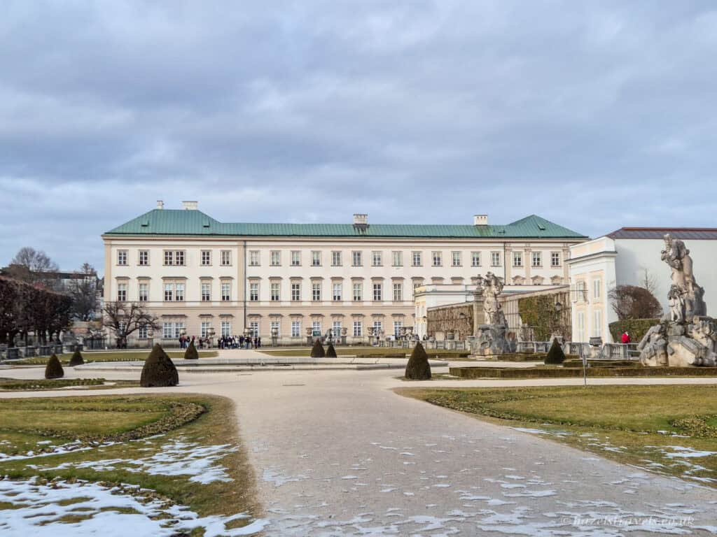Mirabell Palace in Salzburg with pale cream façade and green roof, seen across the formal Mirabell Gardens with statues, gravel paths, and patches of snow under a cloudy sky