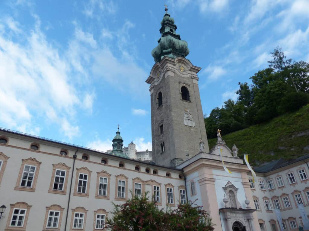 St Peterās Abbey in Salzburg with its tall grey stone tower and green onion dome, set against a blue sky with Hohensalzburg Fortress on the hillside behind