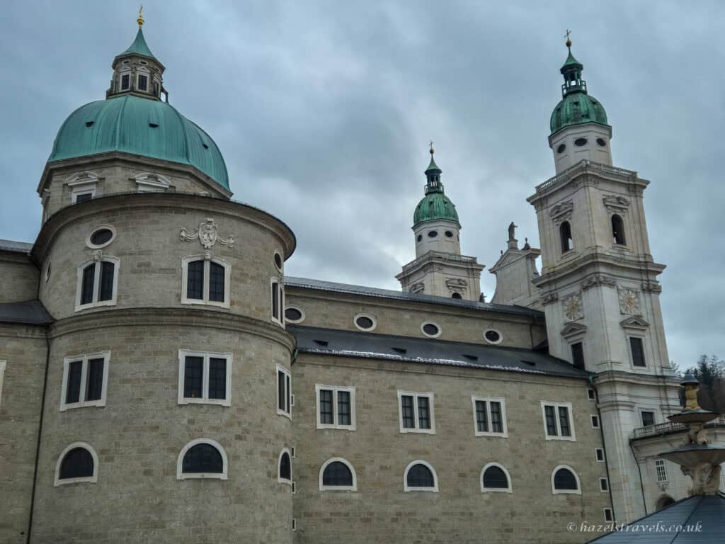 Salzburg Cathedral with large green copper dome and twin towers rising above pale stone walls under a cloudy grey sky